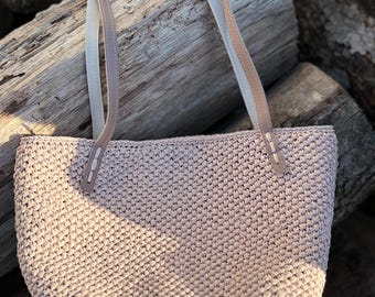May include: A light beige woven tote bag with a rectangular shape. The bag has two handles with a light beige and white color scheme. The bag is made of a woven material and has a textured appearance. The bag is sitting on a pile of wooden logs.