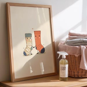 Op de afbeelding: Een ingelijste print met twee cartoon sokken die elkaars hand vasthouden, een gestreept en de andere oranje, met een klein roze hartje erboven. De print zit in een licht houten frame, naast een rieten mand en een fles Lemon Linen spray.