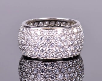 May include: A wide, silver-toned ring encrusted with numerous small, sparkling stones. The ring's surface is completely covered in the stones, creating a brilliant, textured appearance. The ring is reflected in the surface below.