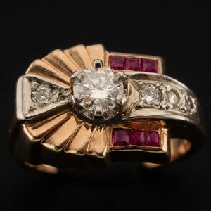 May include: A gold ring featuring a central round brilliant-cut diamond surrounded by a fan-shaped setting. The band includes smaller round diamonds and two rows of three square-cut red rubies set into the polished gold metal surface.