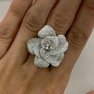 May include: A large, silver-toned ring in the shape of a flower, set with numerous small, sparkling stones. The ring's design features multiple petals, creating a three-dimensional floral effect. The ring is worn on a finger.