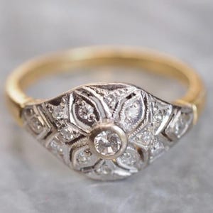 May include: A vintage-style ring with a gold band and a silver-tone setting. The setting features a central round diamond surrounded by smaller diamonds in an ornate, floral-inspired design. The ring is likely made of gold and silver.