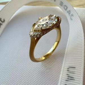 Könnte beinhalten: Ein goldener Verlobungsring mit einem großen, markisenförmigen Diamanten, der von einem Halo aus kleineren Diamanten umgeben ist.