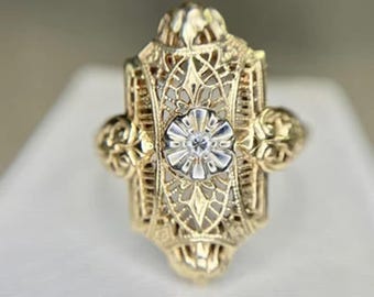 May include: A gold filigree ring with a central floral design and a small, clear gemstone. The ring has an ornate, openwork design with decorative elements on the sides. The ring is set against a neutral background.