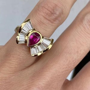 May include: A gold ring featuring a pear-shaped, deep red gemstone at the center, surrounded by a fan-like arrangement of baguette-cut, clear gemstones. The ring's design includes a gold band and setting, creating a luxurious and elegant appearance.