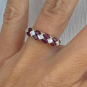 Op de afbeelding: Een gouden ring met een reeks ovaal geslepen, dieprode edelstenen afgewisseld met vierkante, heldere edelstenen. De ring wordt op een vinger gedragen, wat het ingewikkelde ontwerp en de levendige kleuren laat zien.