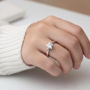 May include: A silver ring with a star-shaped, clear gemstone. The ring is on a person's finger, with the hand in a relaxed position. The ring's band is thin and polished, and the star-shaped stone is the focal point.