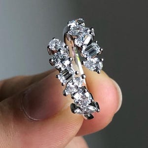Può includere: Un anello d'argento con numerose pietre preziose chiare e scintillanti. L'anello presenta un gruppo di pietre rotonde e a taglio baguette, creando un effetto abbagliante. Le pietre sono incastonate in una fascia curva e delicata.
