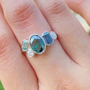 May include: A silver ring featuring an oval-cut teal gemstone, flanked by emerald-cut blue and teal stones, and two small round white gemstones. The ring is set on a hand, showcasing the intricate design and color variations of the gemstones.