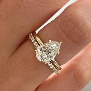 May include: A gold engagement ring with a large, pear-shaped diamond as the centerpiece. A smaller band with embedded diamonds sits next to it. The rings are on a finger, showcasing the jewelry's sparkle and design.