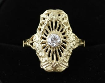 May include: A gold-colored ring with an ornate, filigree design. The ring features a central, round, clear gemstone. The ring's design includes a sunburst pattern around the gemstone and floral accents. The ring is set against a black background.