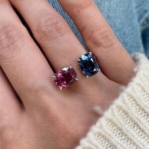 May include: Two gemstone rings, one with a deep pink stone and the other with a blue stone, are displayed. The rings have a simple, elegant design, with the stones set in a silver metal. The rings are worn on a finger.