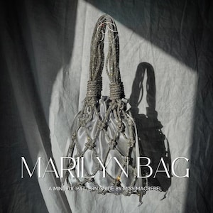 May include: A silver-colored handbag with a drawstring closure and a macrame-style net overlay. The bag has two handles made of a beaded material. The text "MARILYN BAG" is displayed on the front, along with the text "A MINDFUL PATTERN GUIDE BY MISS MACREBEL".