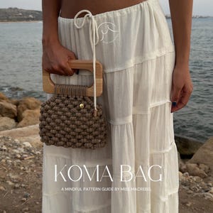 May include: A handcrafted macrame bag with a wooden handle and a gold-colored clasp. The bag is a neutral brown color and is held by a person wearing a white skirt. The text "KOMA BAG" is displayed below the bag.