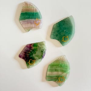 May include: Four fluorite gua sha tools. Each tool is a different shade of green, purple, and white. A gold heart design is on each tool. The tools are on a white background.