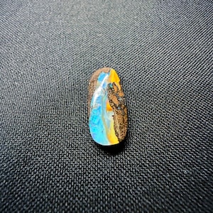 Könnte beinhalten: Ein tropfenförmiger Boulder-Opal-Edelstein mit einer polierten Oberfläche, die lebhafte Blitze in Elektroblau und hellem Gelb aufweist, kontrastiert durch eine texturierte, dunkelbraune Eisensteinmatrix an der Seite und Oberseite.