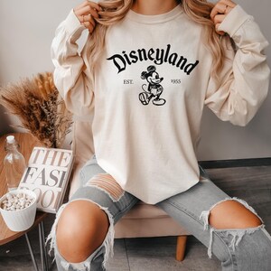 May include: A cream-colored long-sleeved sweatshirt with a black graphic of Mickey Mouse and the text "Disneyland Est. 1955".