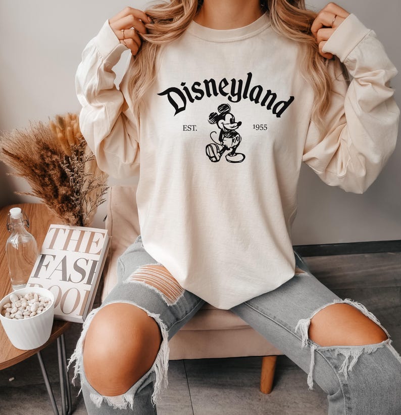 May include: A cream-colored long-sleeved sweatshirt with a black graphic of Mickey Mouse and the text "Disneyland Est. 1955".