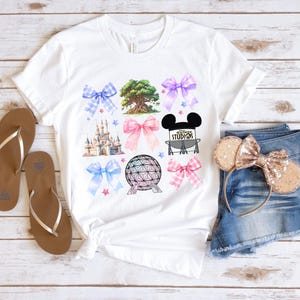 May include: White t-shirt with a colorful design featuring a castle, a tree, a bow, a Mickey Mouse head, a park icon, and a sphere. The design is surrounded by pink, blue, and white bows.