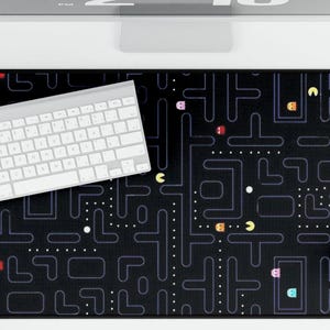 May include: Black rectangular desk mat featuring a Pac-Man maze design with colorful ghosts and Pac-Man characters. A white keyboard and mouse are on the mat. The mat is 30 inches long and 12 inches wide.