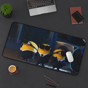 May include: A rectangular black desk mat featuring three Minions from the Despicable Me franchise, wearing sunglasses and suits. The background shows a car and a street scene. A laptop, mouse, and coffee cup are also visible.