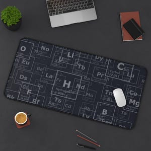 May include: A black rectangular desk mat with a periodic table design in white. A white computer mouse, a laptop, a small potted plant, a cup of coffee, a notebook, and writing utensils are on the dark surface.