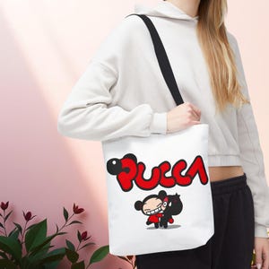 May include: White tote bag featuring the word "Pucca" in red and black, with a cartoon illustration of two characters. The bag has a black shoulder strap and is held by a person.