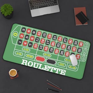 May include: A green roulette-themed desk mat with a white mouse, a laptop, and a cup of coffee. The mat features a roulette wheel layout with numbers and betting sections. A small potted plant, a notebook, and pens are also on the dark surface.