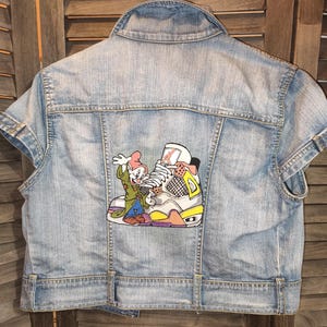 Peut inclure: Veste en jean bleu clair avec un motif de dessin animé dans le dos. Le motif représente un personnage de dessin animé et une basket colorée. La veste a des manches courtes et une fermeture boutonnée.