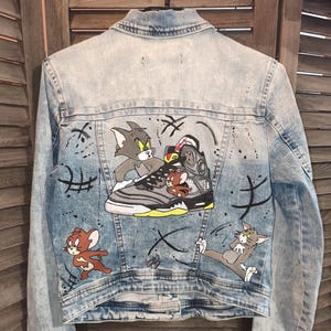 Può includere: Giacca di jeans azzurra con un design a cartoni animati con i personaggi di Tom e Jerry. La parte posteriore della giacca presenta un design di sneaker con i personaggi dei cartoni animati e macchie di vernice nera.