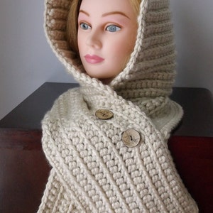 May include: A cream-colored, hooded scarf made of thick, textured yarn. The scarf features a ribbed pattern and fastens with two wooden buttons. The hood is designed to frame the face. A cozy winter accessory.