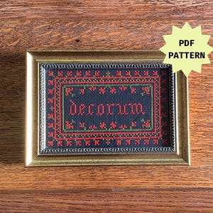 May include: A framed cross-stitch embroidery with a black background and red and green accents. The embroidery reads "decorum" in a gothic font.