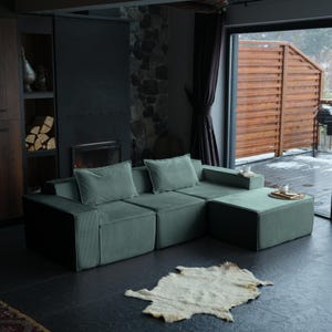 May include: A modern, modular sectional sofa in a muted green hue, with two matching square throw pillows. The sofa is paired with a light-colored, textured rug. The room features a fireplace, wooden accents, and a view of an outdoor deck.
