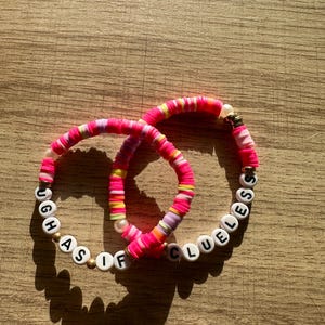 May include: Two pink and white beaded bracelets with the words "Ugh As If" and "Clueless" spelled out in black letters.