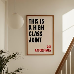 May include: A rectangular wall art print in a thin brown frame featuring the black text "THIS IS A HIGH CLASS JOINT" and the red text "ACT ACCORDINGLY" on a cream background. The print is displayed on a plain wall next to a staircase.