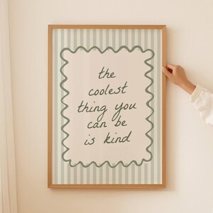 May include: A rectangular framed wall art piece featuring a light green and white vertical striped background. Centered is a cream-colored rectangle with a wavy dark green border containing the dark green text: the coolest thing you can be is kind.