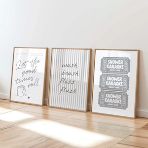 May include: Three framed wall art prints in light wood frames. The first print features a guest check design with the text Let the good times roll and a toilet paper roll. The second has vertical stripes and the words wash brush floss flush. The third shows three grey tickets labelled SHOWER KARAOKE.