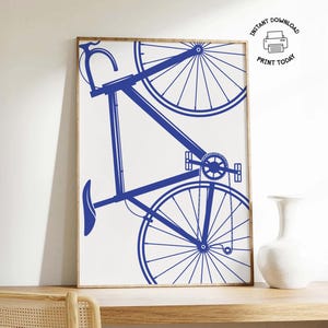 Op de afbeelding: Een ingelijste print met een blauwe fietsillustratie op een witte achtergrond. De fiets is afgebeeld in een minimalistische stijl, met de wielen en het frame duidelijk omlijnd. De print wordt weergegeven op een houten oppervlak, met een witte vaas rechts. De tekst "INSTANT DOWNLOAD PRINT TODAY" is zichtbaar.