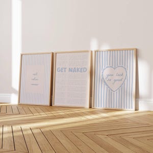 May include: Three rectangular wall art prints in light wooden frames. The left print has blue stripes and the text rest relax renew. The center print features the repeated text GET NAKED. The right print has blue stripes and a heart with you look so good.