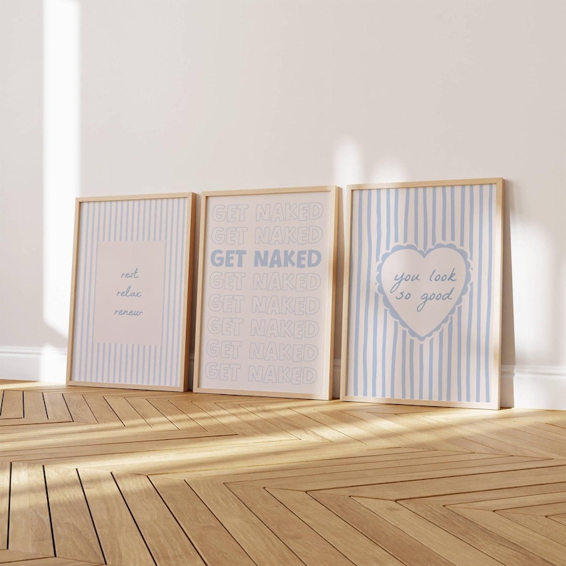 May include: Three rectangular wall art prints in light wooden frames. The left print has blue stripes and the text rest relax renew. The center print features the repeated text GET NAKED. The right print has blue stripes and a heart with you look so good.