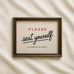 Pode incluir: Uma placa retangular numa moldura de bronze escuro com fundo creme. O texto diz PLEASE em vermelho, seguido de seat yourself em letra cursiva preta, e NO RESERVATION NEEDED em letras maiúsculas pretas pequenas na parte inferior.