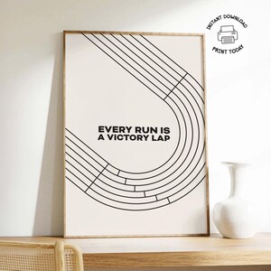 Puede incluir: Una impresión enmarcada con el texto "EVERY RUN IS A VICTORY LAP" en negro. El diseño presenta líneas negras que forman una forma de pista sobre un fondo crema. Un jarrón blanco está sobre una superficie de madera.