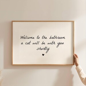 Könnte beinhalten: Ein rechteckiges Wandbild in einem hellen Holzrahmen mit dem schwarzen Text "Welcome to the bathroom a cat will be with you shortly", zentriert auf einem cremefarbenen Hintergrund mit einem kleinen schwarzen Herzsymbol unter dem Text.
