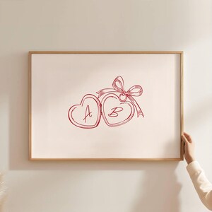 May include: A framed print featuring a red line drawing of two heart-shaped lockets, each with a letter inside, connected by a ribbon and bow. The letters "A" and "B" are visible. The frame is a light brown color.