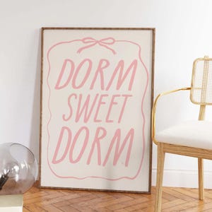 May include: A rectangular wall art print in a wooden frame featuring a cream-colored background with a pink hand-drawn border and a bow at the top. The text DORM SWEET DORM is written in large, pink, handwritten-style capital letters in the center.