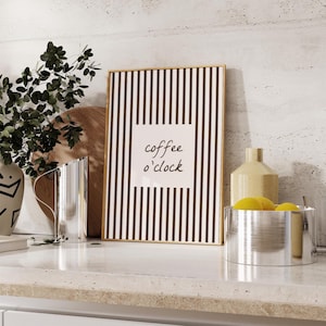 Puede incluir: Un cuadro rectangular con marco que presenta un patrón de rayas verticales blancas y negras con un cuadrado blanco central que contiene el texto manuscrito en negro coffee o'clock. El marco es fino y de color dorado.