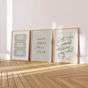 Three sage green and pastel mint printable bathroom wall art posters leaning against a wall, featuring a funny toilet print with “let the good times roll,” a striped wash brush floss flush sign, and a retro shower karaoke ticket-style design, styled as trendy aesthetic restroom decor.