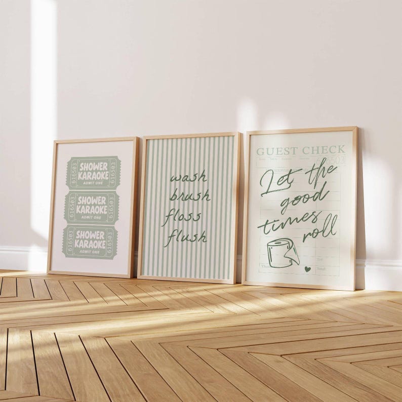 Three sage green and pastel mint printable bathroom wall art posters leaning against a wall, featuring a funny toilet print with “let the good times roll,” a striped wash brush floss flush sign, and a retro shower karaoke ticket-style design, styled as trendy aesthetic restroom decor.
