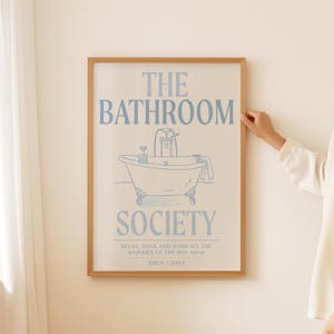 May include: A rectangular wall art print in a light wood frame featuring blue line art of a clawfoot bathtub. The text reads THE BATHROOM SOCIETY, RELAX, SOAK AND WASH ALL THE WORRIES OF THE DAY AWAY, and OPEN 7 DAYS on a cream-colored background.