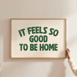 May include: A framed print with the text "IT FEELS SO GOOD TO BE HOME" in green letters on an off-white background. The frame is a light brown color. A person's hand is holding the frame.